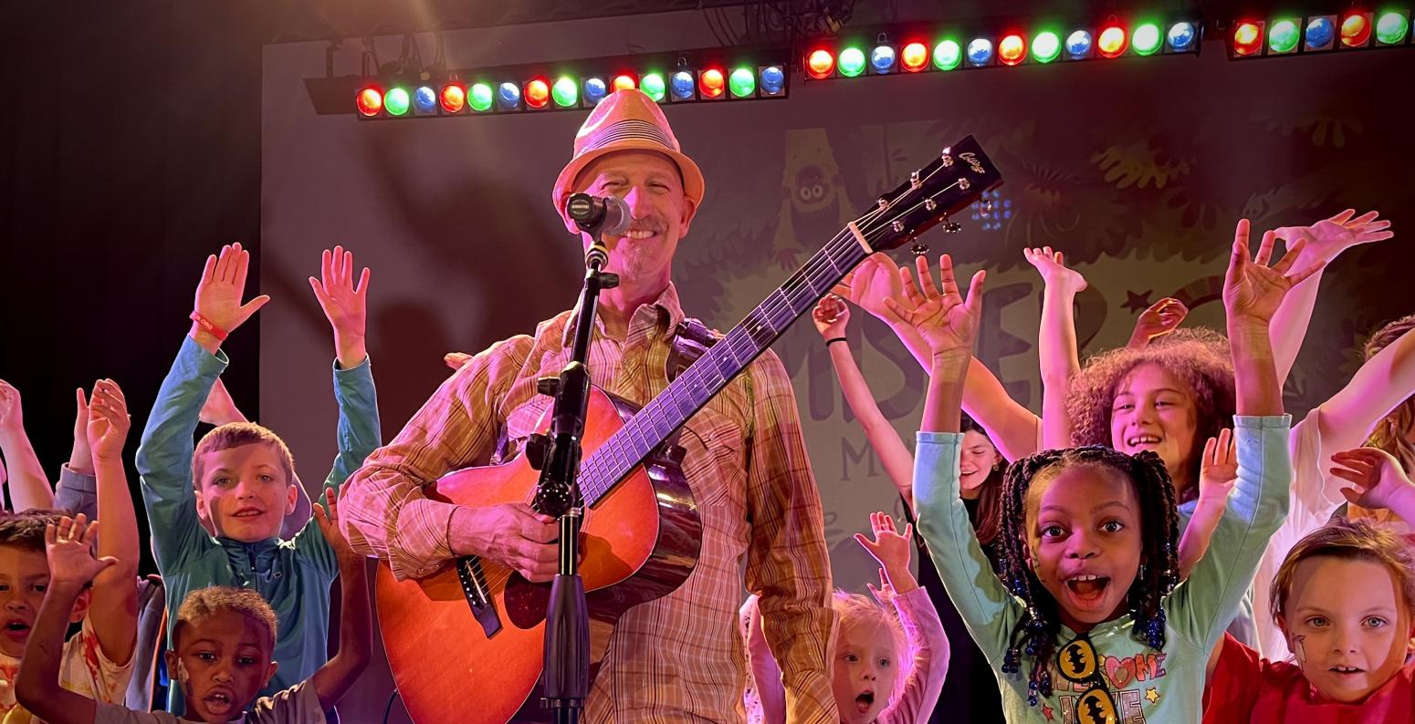 MISTER G standing in front of a microphone holding a guitar surrounded by smiling children with their hands up.