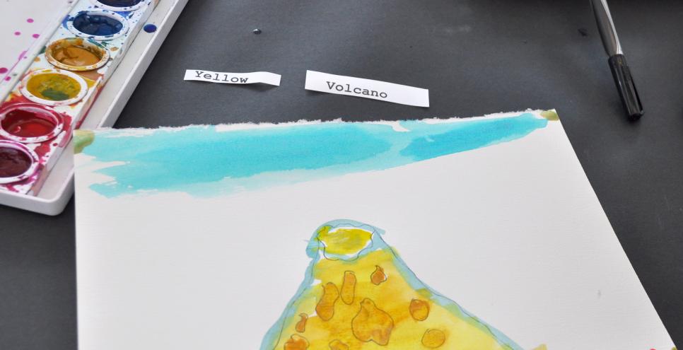 Two paper slips with the words "yellow" and "volcano" next to a watercolor painting of a yellow volcano against a blue sky.