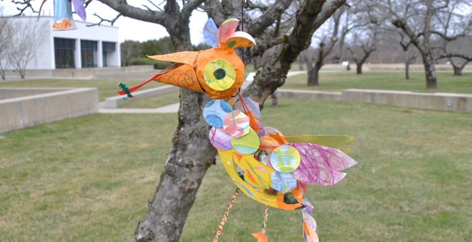 bird mobiles in bobbie's meadow. projects are hanging in a tree. Birds are made out of collaged papers and materials with bright orange, yellow, blue and purple. 
