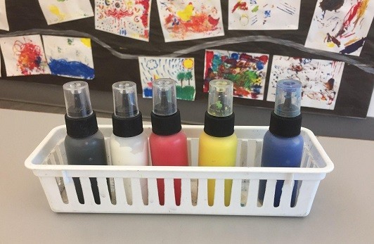 Basket with five 2-ounce bottles filled with paint in front of a wall with paintings displayed.