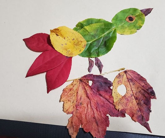 Collage of a bird perched on a branch. The bird is made of bright red, yellow, and green leaves. 