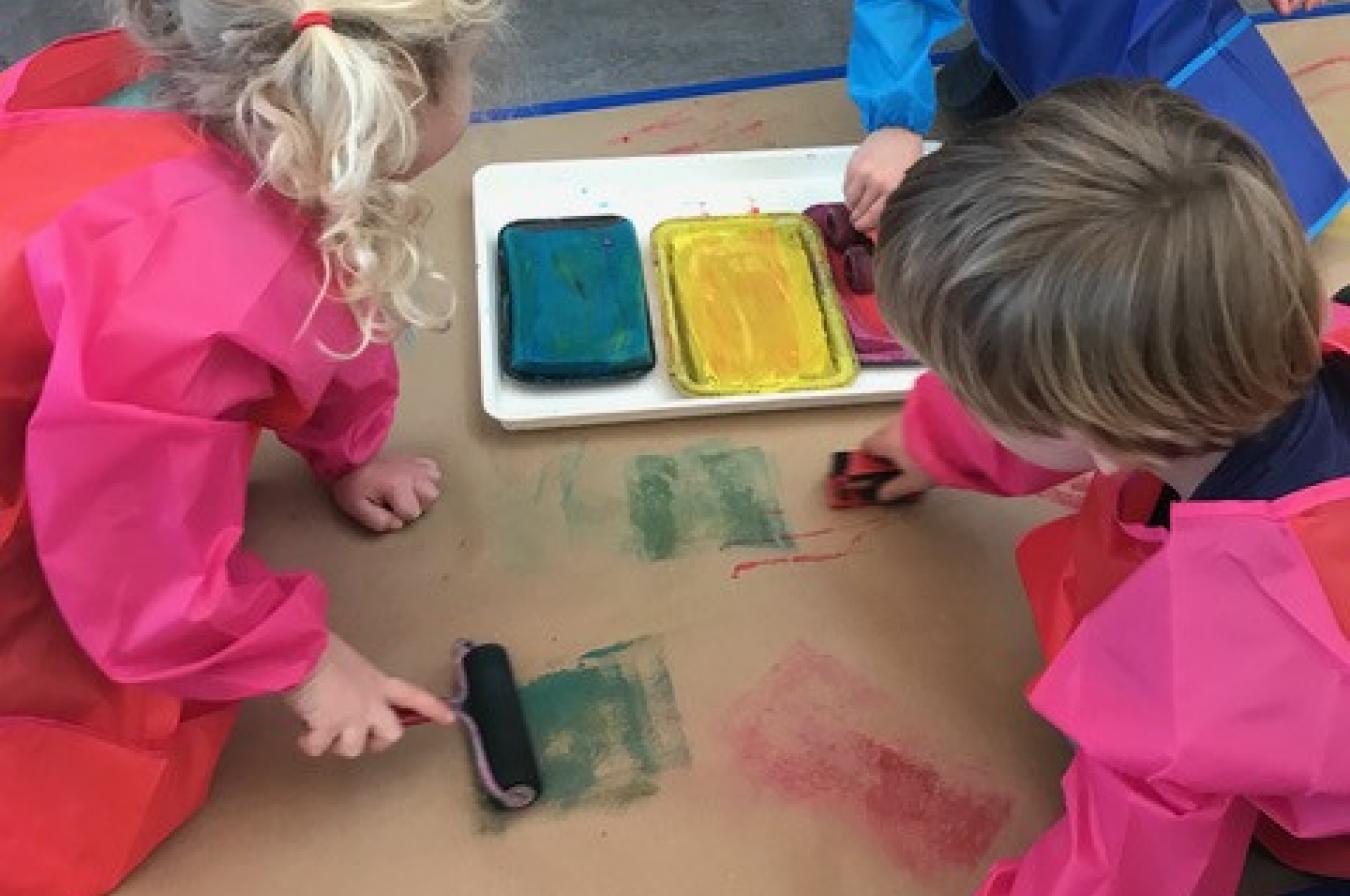 Photo shows three children rolling colored paint on cardboard with paint rollers and toy cars.