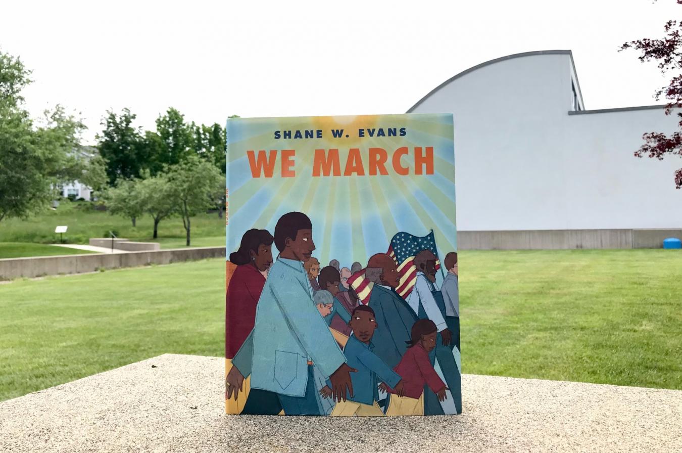 The book We March is propped on an outdoors table. Green grass and The Eric Carle Museum can been seen in the background.