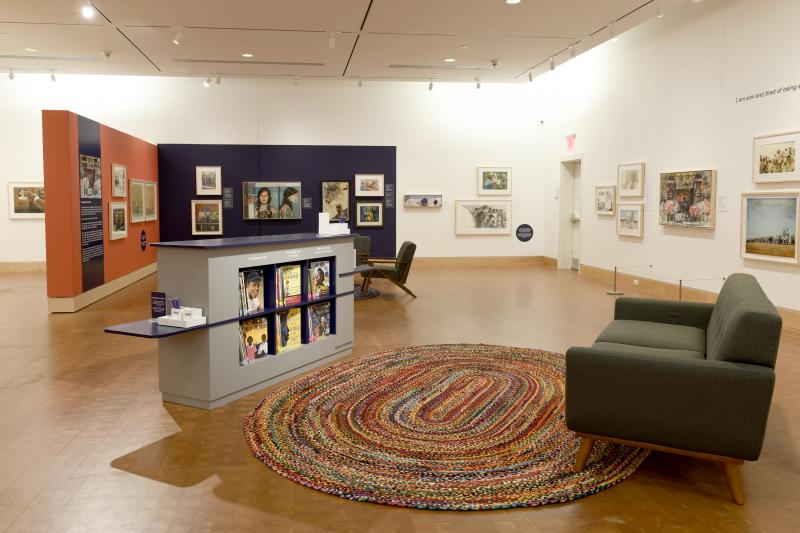 Installation image showing walls with framed art and reading area with sofa and bookcase.