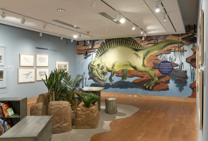 Installation image showing walls with framed art and large Spinosaurus mural.
