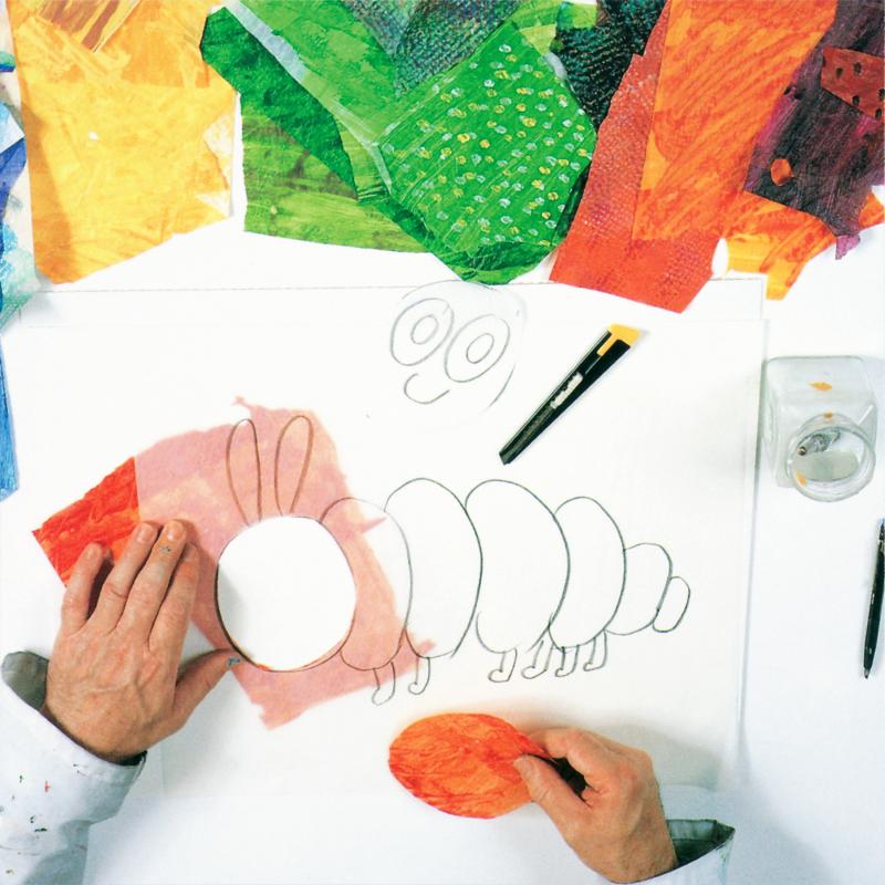 hand holding red painted tissue paper oval cut out and tracing paper with caterpillar drawing missing it's face, which is above on the table