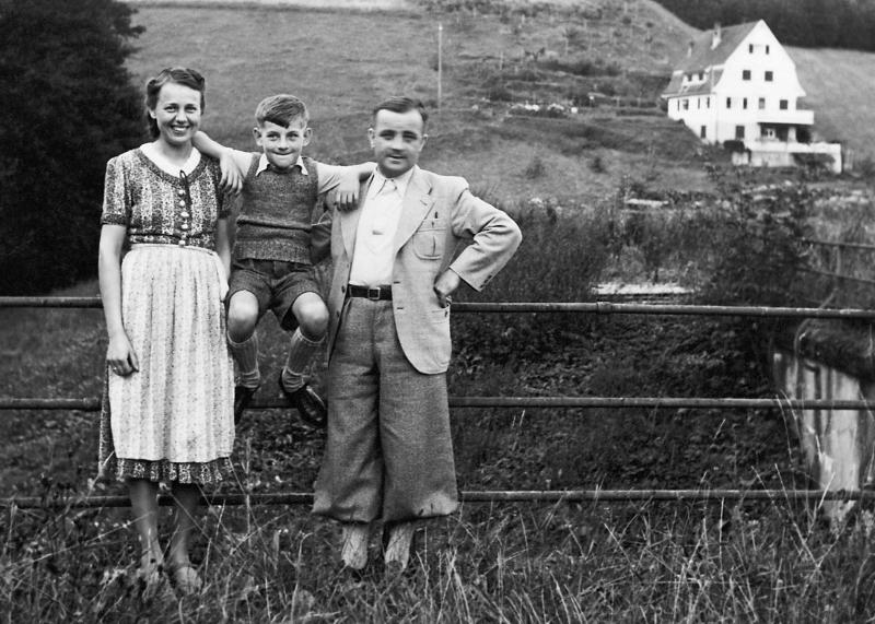 parents with child sitting on a fence with white house in the background mountains