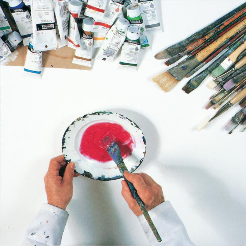 overhead photograph of hands holding a paintbrush in a bowl of red paint