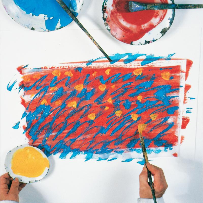 hand with paintbrush adding yellow dots to tissue paper painted red with blue lines