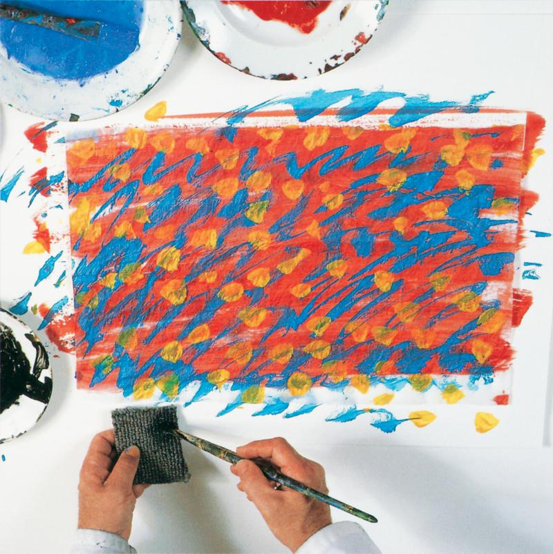 hand with paintbrush adding dark green paint to small carpet square next to painted tissue paper