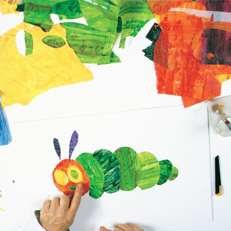hand placing green eye ball tissue paper shape in right yellow tissue paper eye, left eye is still just yellow tissue paper and green nose is now on red oval caterpillar face