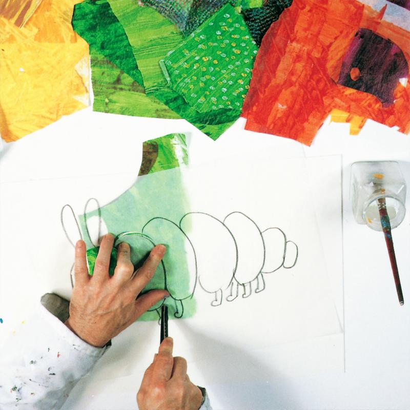 hand holding tracing paper with caterpillar drawing above green painted tissue while second hand cuts with a utility blade.