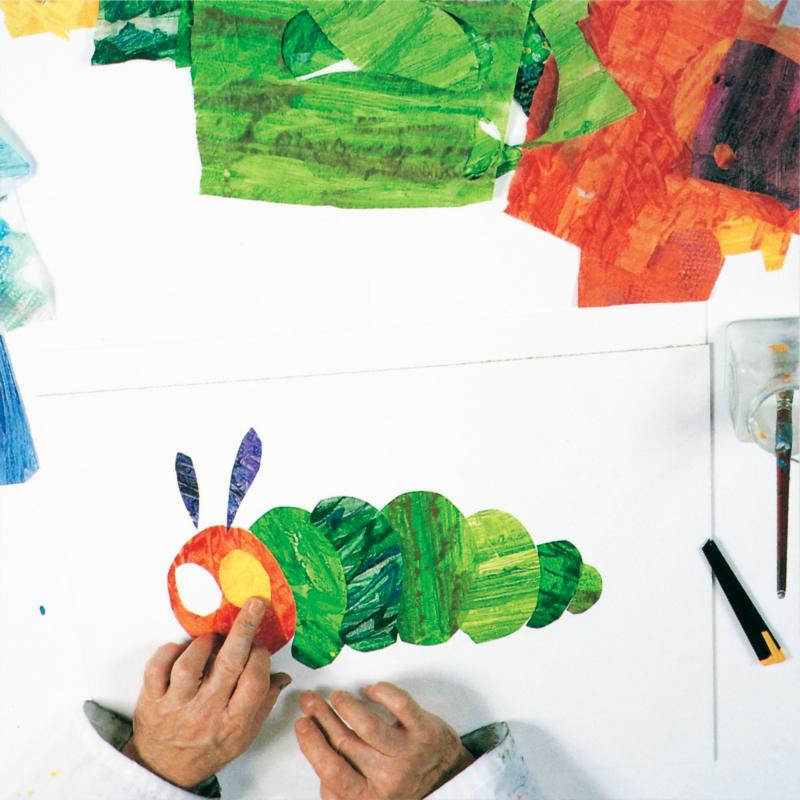 hand placing yellow painted tissue paper eye on red oval tissue paper. The caterpillar now has two blue antennas and six green shapes made from different green painted tissue papers