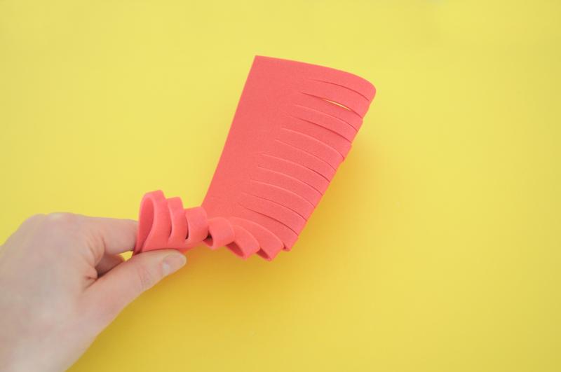 A hand folds a pink piece of thin foam with cuts made along the folded edge of the foam.