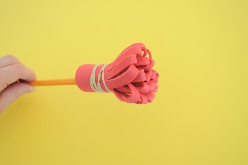 A hand holding a homemade brush, with the pink foam attached to the end of a pencil with a rubber band.