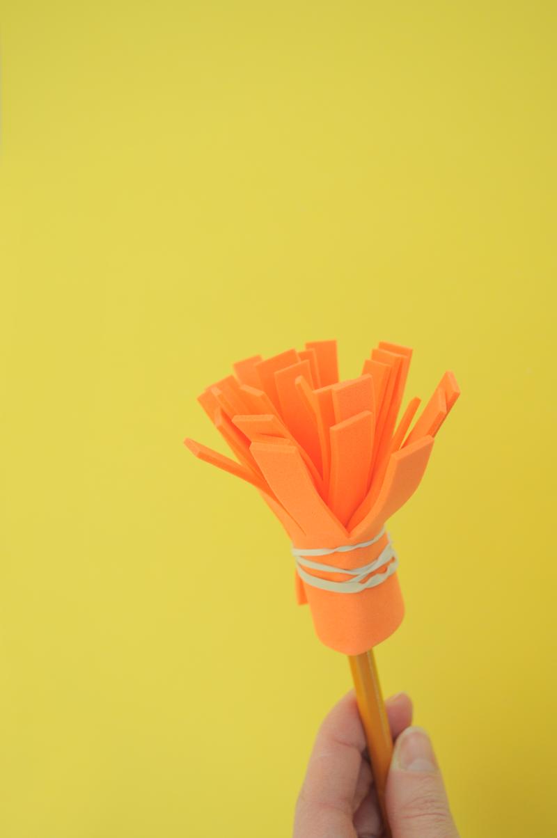 A hand holding a homemade brush, with the orange foam attached to the end of a pencil with a rubber band.
