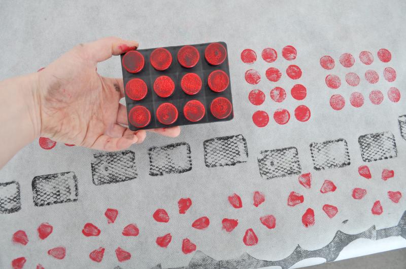A hand holds a black plastic block with circles on it covered in red paint.