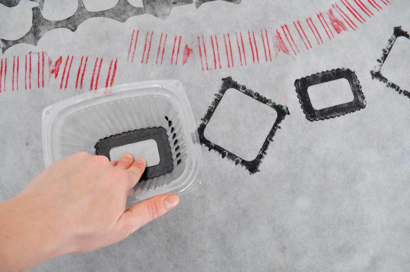 A hand presses a plastic container covered in black paint onto a white surface.