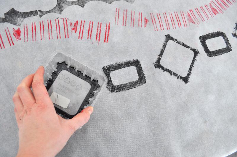 A hand presses a plastic container covered in black paint onto a white surface making a diamond shape.