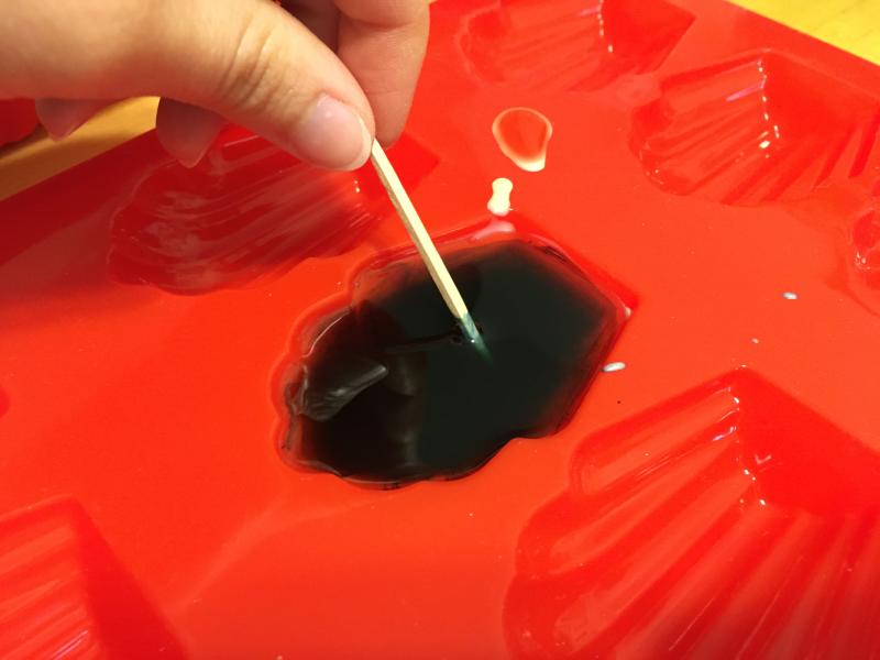A hand mixes blue colorant into the melted wax with a toothpick.