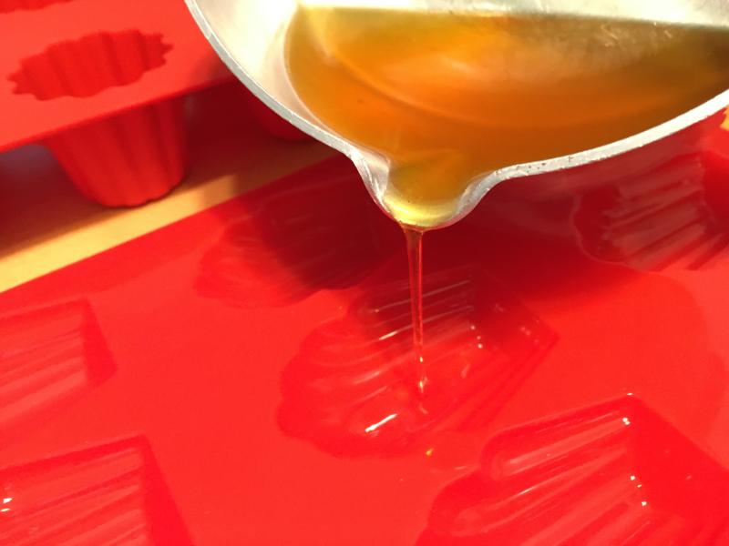A stream of wax pouring out of a silver pot into a bright red silicone mold.