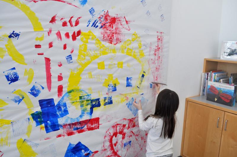 A young child paints on a large mural wall with a piece of cardboard dipped in paint.