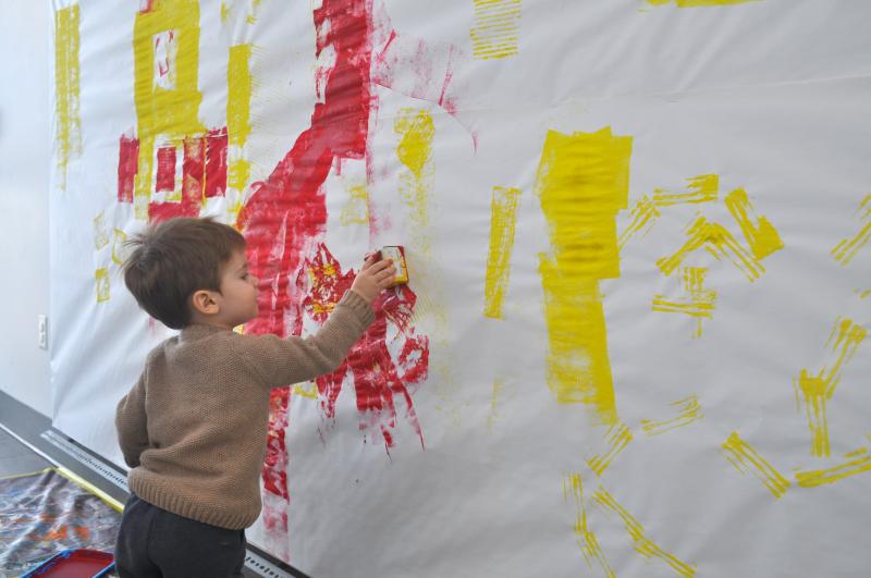 A young child presses a box dipped in paint onto a large mural wall.