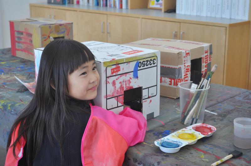 A young child smiles while their hand is hidden inside of a box, painting with watercolor.