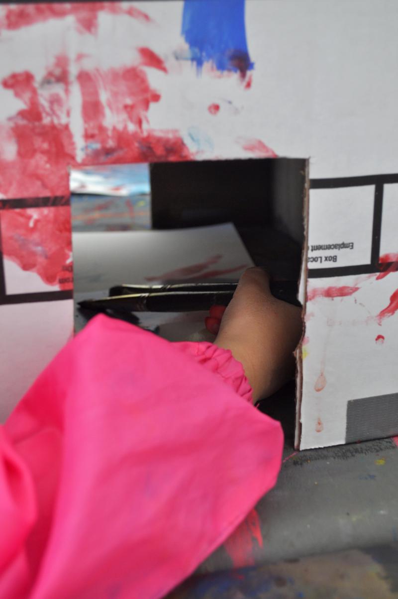 A hand paints with two paintbrushes inside of a box.