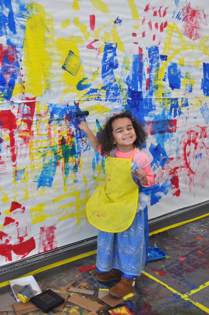 A young child smiles while holding a painty brayer up to a large mural painting.