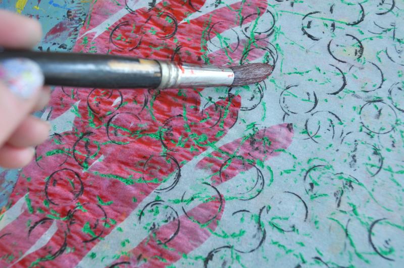 A brush paints on watercolors over crayon rubbings.