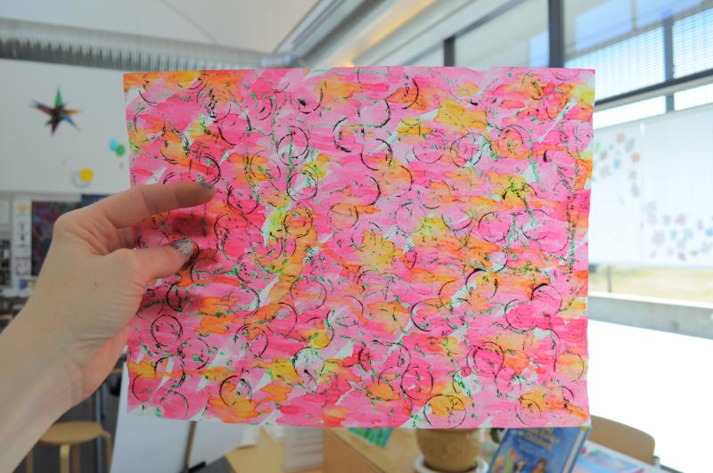 A hand holds up a watercolor-painted paper to the sun, allowing light to shine through the translucent paper.