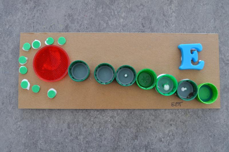 An abstract caterpillar mixed-media collage with green and red plastic caps on brown cardboard.