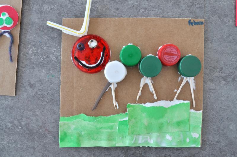 A caterpillar artwork with a straw for antennae, body made from bottle caps, and paper grass.