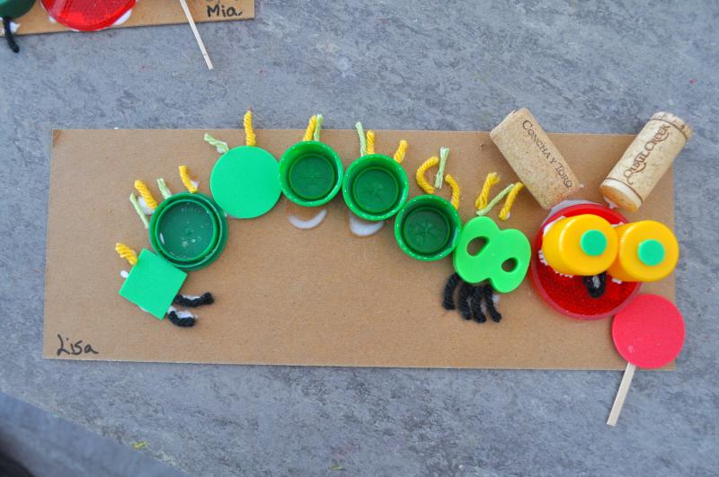 A mixed-media collage of a caterpillar created with yarn, plastic caps, and cork, with a paper lollipop in front of it.