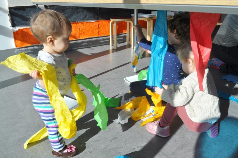 A young guest holds long strips of tissue paper in their hands as they watch two older visitors tear tissue papers from underneath a table.
