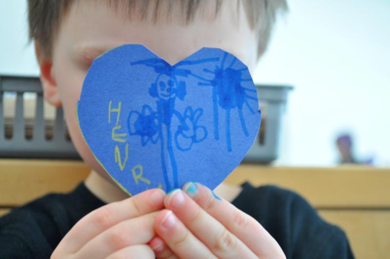 A child holds up a blue paper heart with a written name on it and a drawing of a person.
