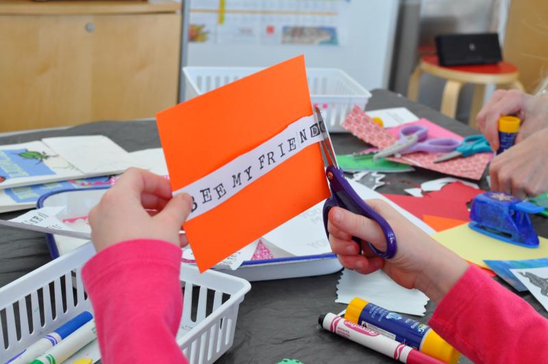 A child cuts a printed phrase that says &quot;Bee my friend&quot; and attaches it to their valentine card.