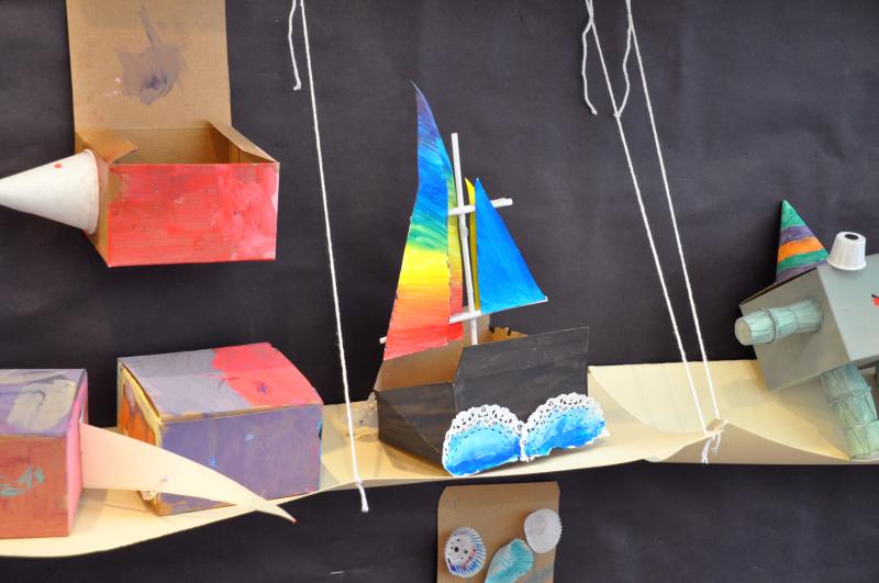 A sculpture of a sailboat with a rainbow sail, a folded box for the bow, and crashing waves made from paper doilies.