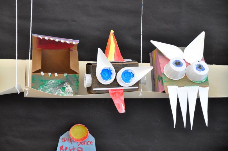 Three sculptures sitting on a paper shelf with prominent teeth, a party hat, cups for eyes, and bagasse product eyebrows.