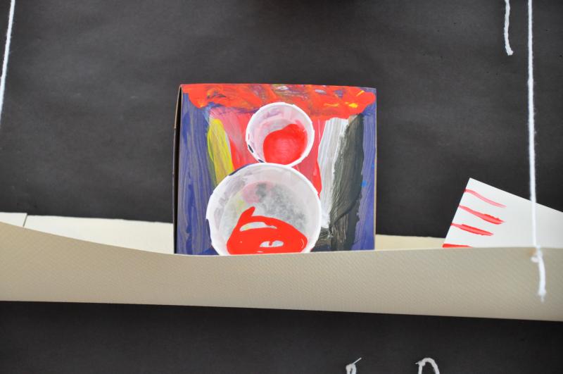 A sculpture sitting on a paper shelf with two cups of different sizes glued to the front and stripes of colorful paint.