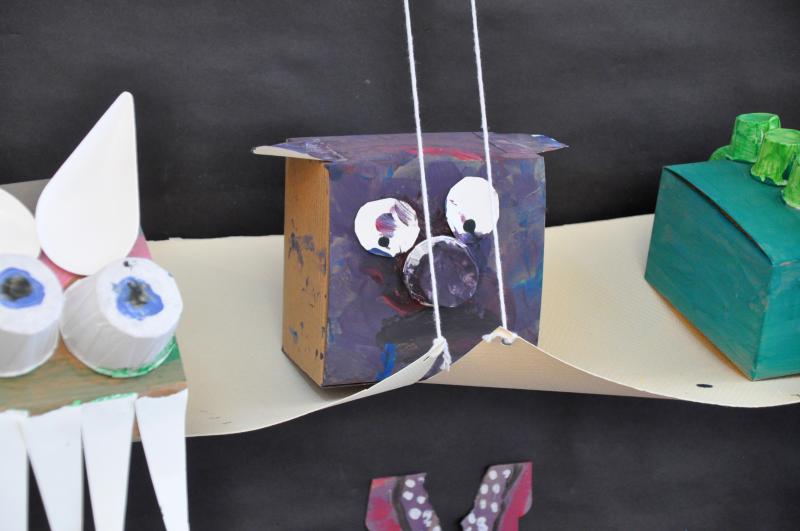 Three sculptures sitting on a paper shelf, one of them with purple colors, torn paper eyes, and a cup for a nose.