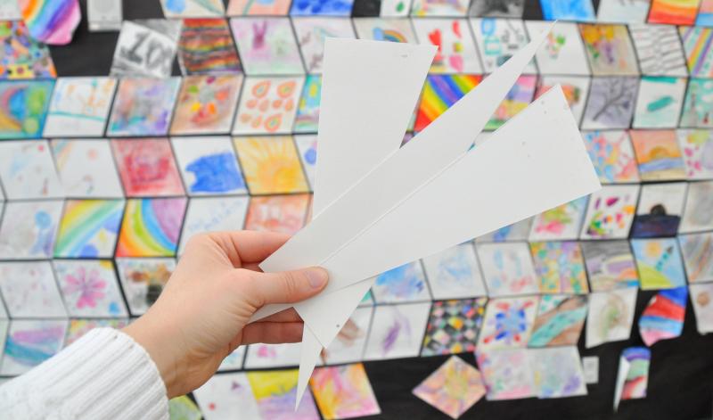 A hand holds up triangular paper offcuts in front of a display of colorful, diamond-shaped artworks.