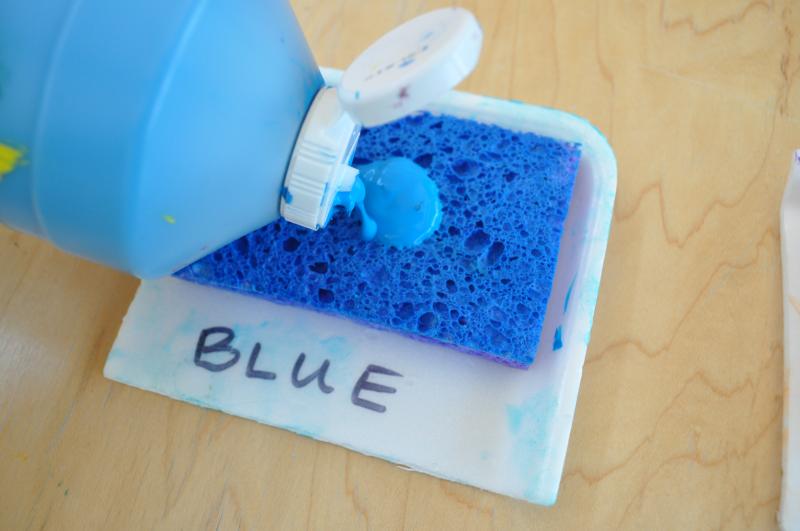 Blue tempera paint being poured onto a stamp pad. It is made of sponge glued onto a plastic tray and labeled “blue.”