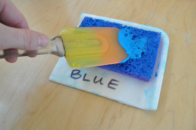 A hand spreading blue tempera paint on a stamp pad using a spatula.