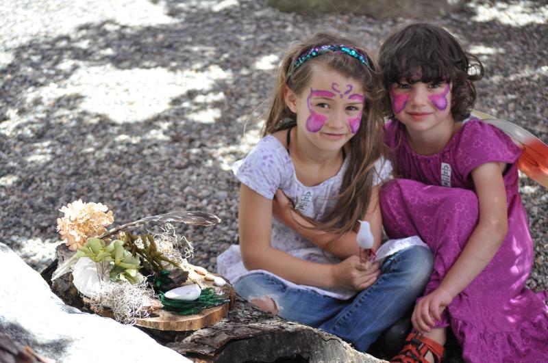 Two young children sit and smile outside near their handmade fairy homes.
