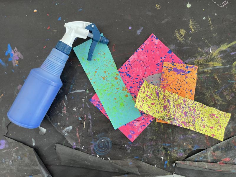 Spray bottle full of watered down paint, bright colorful papers with paint splatter on them.