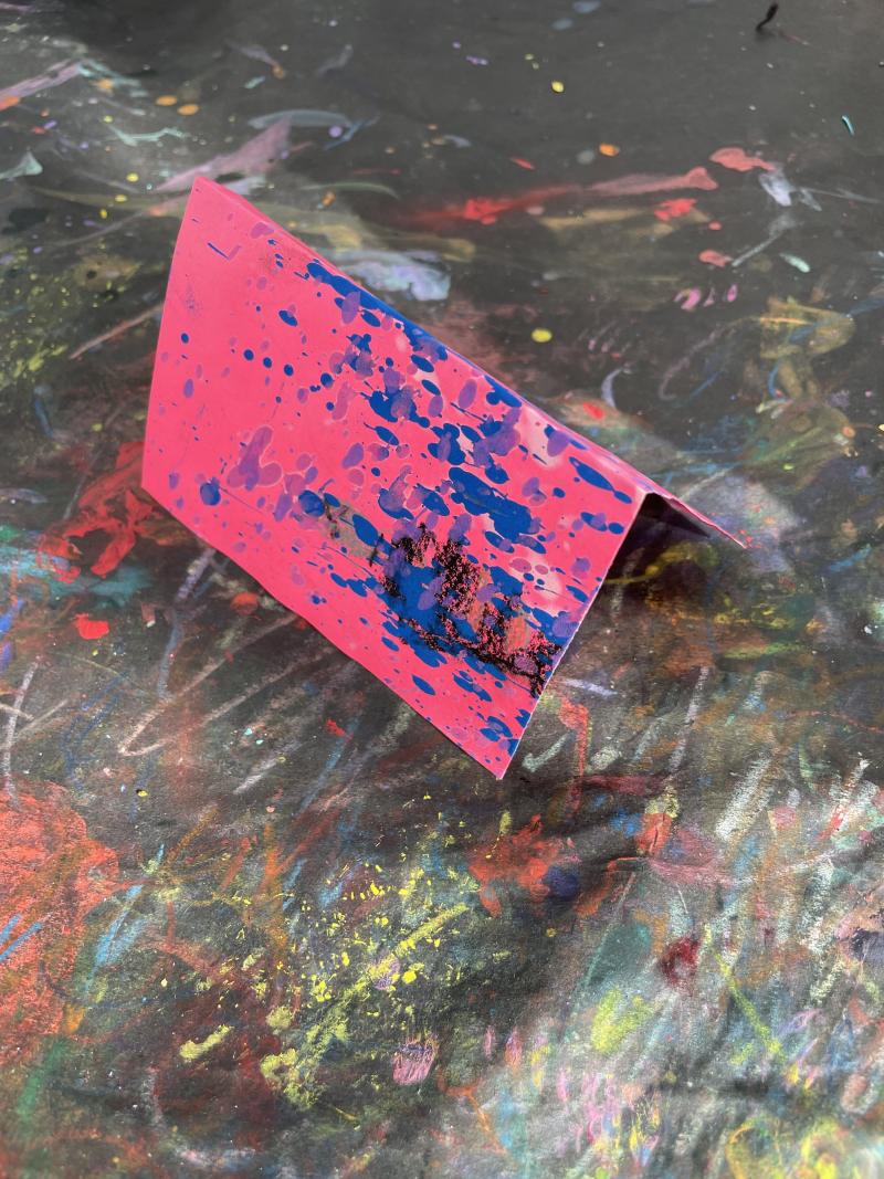 Bright pink paint splattered paper folded in half, standing on table.