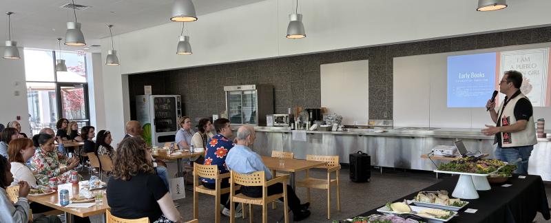 Dr. Lee Francis with a microphone and slides in the background presents to a group in the Museum cafe
