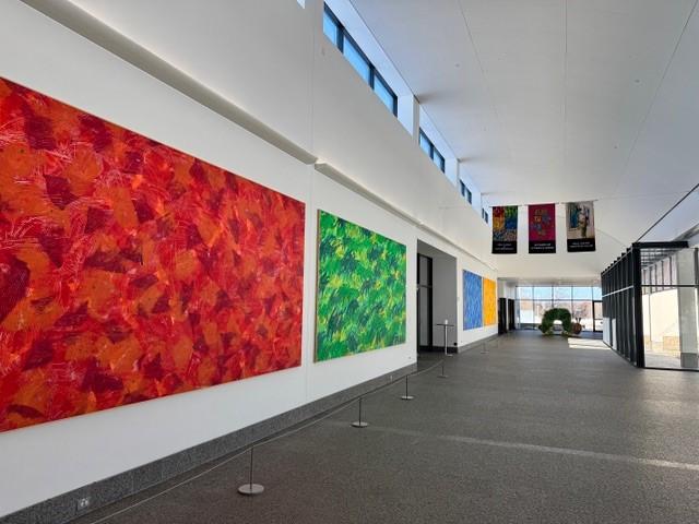 large hall with gray granite floor, big red, green, blue, and yellow murals along the left wall and windows to the right, banners, and a caterpillar display a the end of the hall